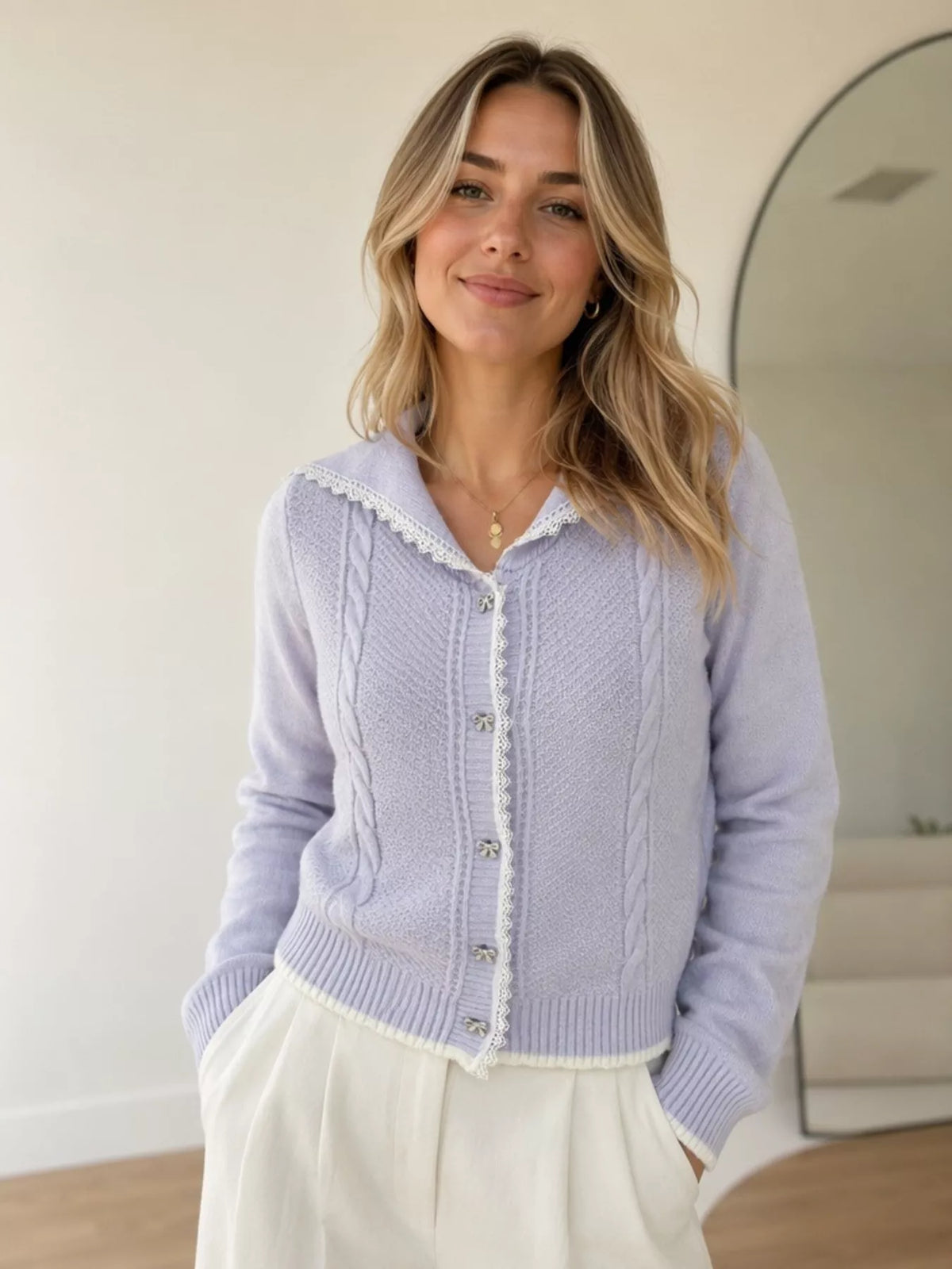 Model wearing a lavender cable-knit cardigan with white lace trim and cream trousers.